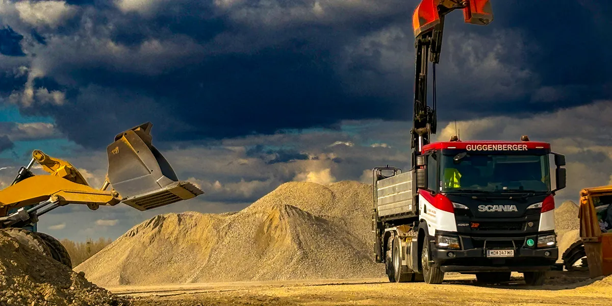 Ein Bagger führt Baggerarbeiten aus, indem er unter einem bewölkten Himmel auf einer Baustelle Kies auf die Ladefläche eines Scania-LKW lädt.